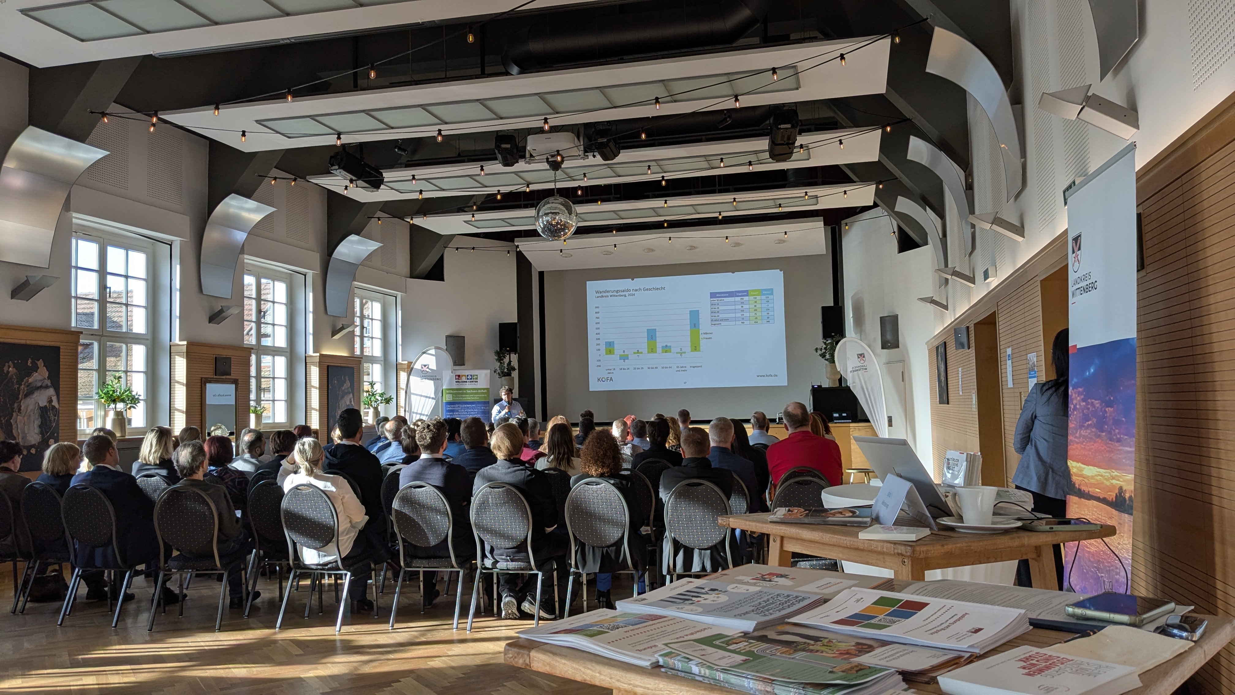 Publikum eines Vortrags von hinten, im Vordergrund ein Infostand mit Flyern und Broschüren