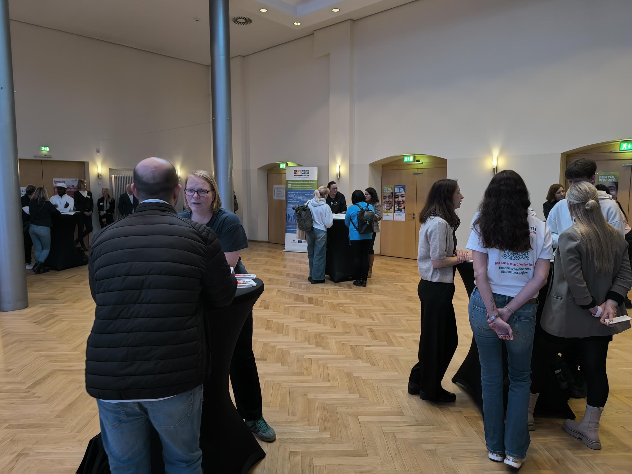 ein Raum mit mehreren Stehtischen, an denen sich kleine Gruppen unterhalten