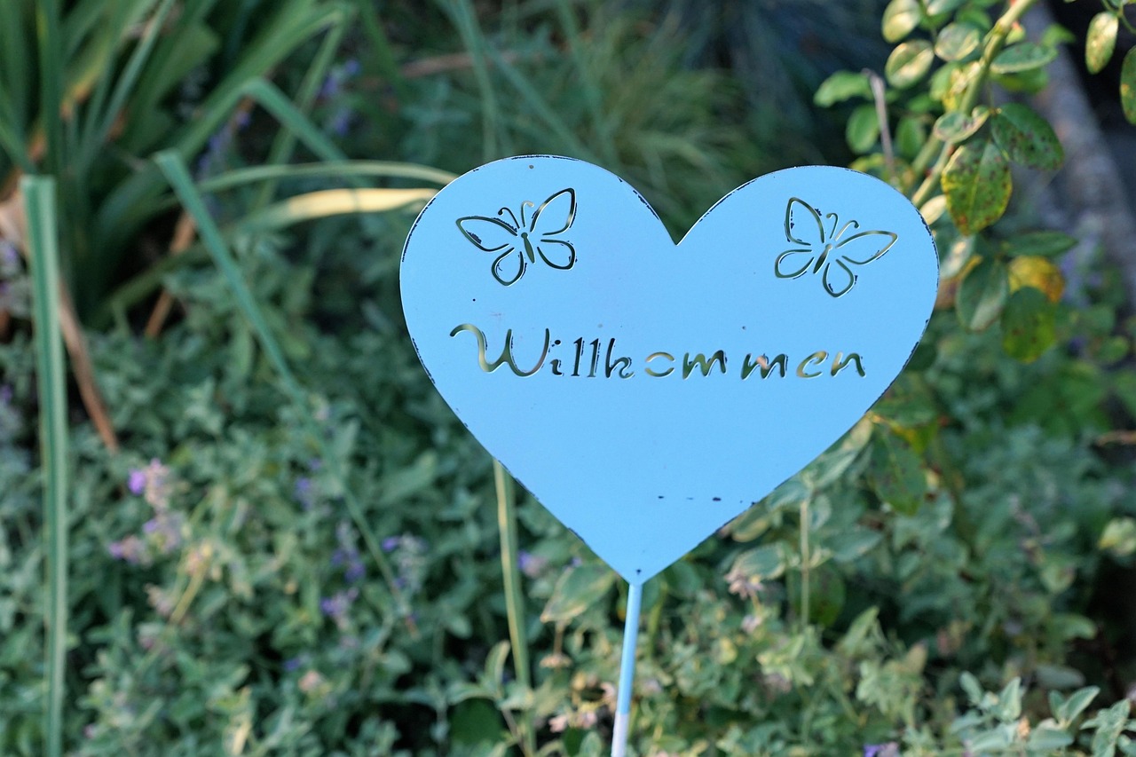 blaues Metallschild in Herzform mit der Aufschrift "Willkommen" und dem Umriss von zwei Schmetterlingen, im Hintergrund ein Garten