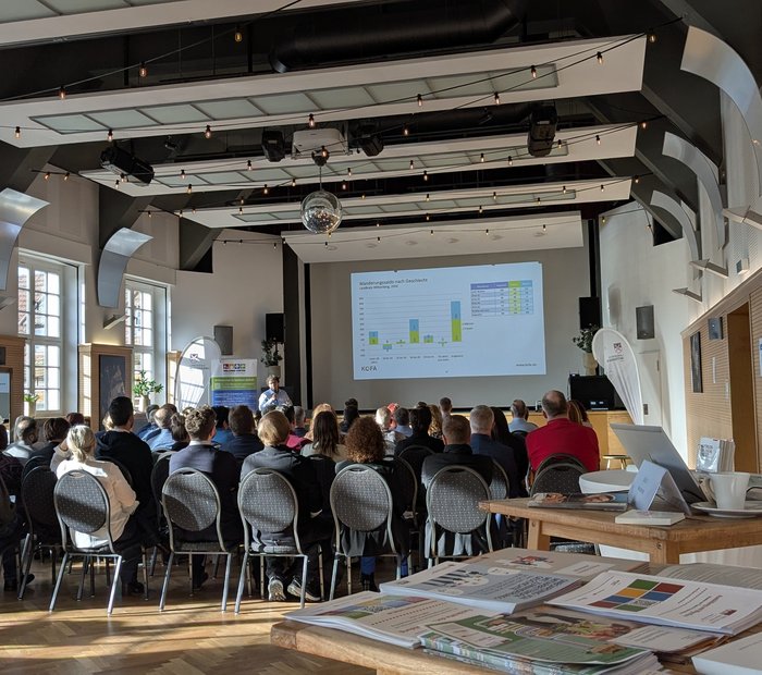 Publikum eines Vortrags von hinten, im Vordergrund ein Infostand mit Flyern und Broschüren