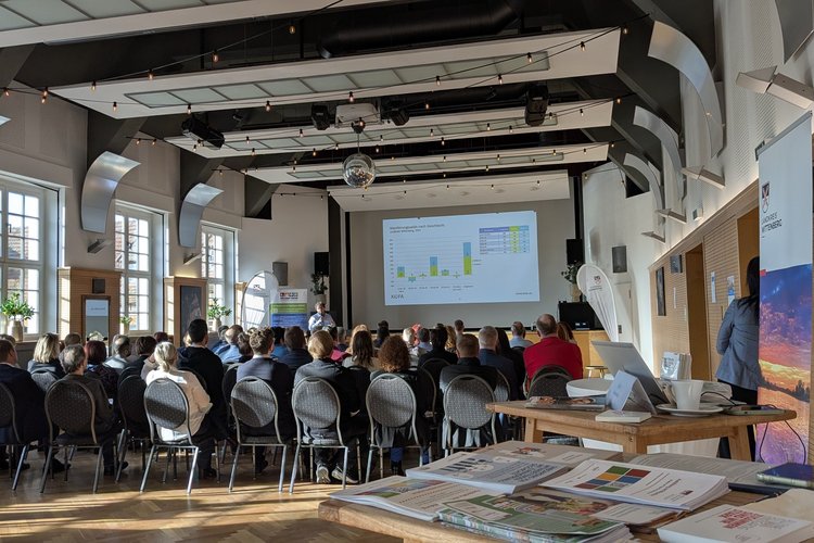 Publikum eines Vortrags von hinten, im Vordergrund ein Infostand mit Flyern und Broschüren