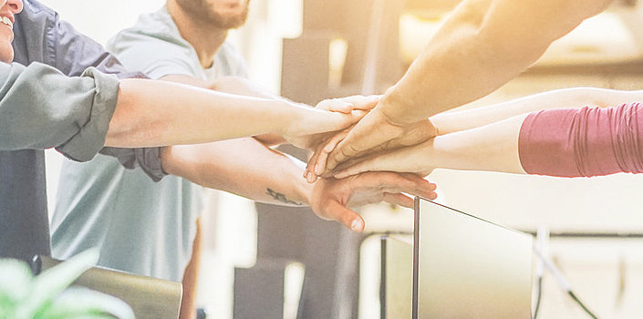 Eine Gruppe von Menschen stehen im Kreis und halten ihre Hände in der Mitte aufeinander. Teamgefühl wird signalisiert.