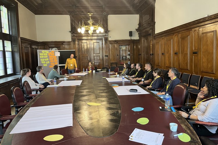 12 Personen sitzen bzw. stehen um einen großen Tisch in einem holzvertäfelten Raum, auf dem Tisch liegen Workshopmaterialien, daneben steht eine mit bunten Zetteln bestückte Moderationswand