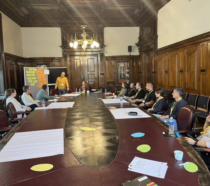 12 Personen sitzen bzw. stehen um einen großen Tisch in einem holzvertäfelten Raum, auf dem Tisch liegen Workshopmaterialien, daneben steht eine mit bunten Zetteln bestückte Moderationswand