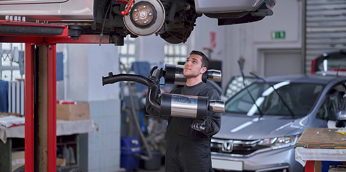 Ein junger Mann steht in Arbeitskleidung mit einem Auspuff unter einem hochgehobenen Auto und schaut hoch, um den Auspuff anubringen