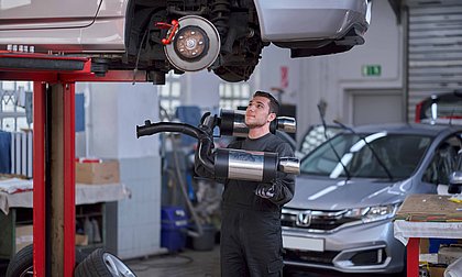 Ein junger Mann steht in Arbeitskleidung mit einem Auspuff unter einem hochgehobenen Auto und schaut hoch, um den Auspuff anubringen