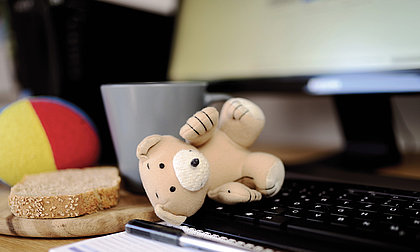 Bild von einem Tisch, auf dem eine Tastatur, ein Butterbrot, eine Kaffeetasse und Kinderspielzeug liegen.