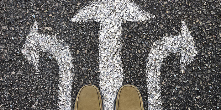 Schuhe von einer männlichen Person auf dem Asphalt, dort sind mit Kreide drei Pfeile aufgemalt. Einer nach links, nach rechts und nach vorne 