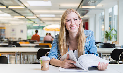 Lächelnde Frau sitzt mit aufgeschlagenem Buch und Kaffeebecher an einem Tisch