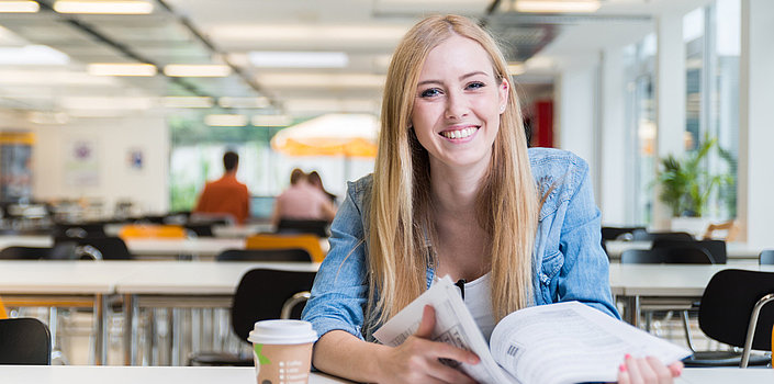 Lächelnde Frau sitzt mit aufgeschlagenem Buch und Kaffeebecher an einem Tisch