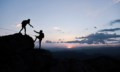 Eine Person steht auf einem Berg und reicht einer anderen Person die Hand, um ihr auf den Berg hoch zu helfen. Im Hintergrund ein schöner Sonnenuntergang.