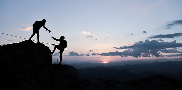 Eine Person steht auf einem Berg und reicht einer anderen Person die Hand, um ihr auf den Berg hoch zu helfen. Im Hintergrund ein schöner Sonnenuntergang.