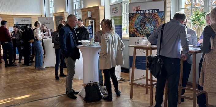 sich unterhaltende Menschen in einem lichtdurchfluteten Veranstaltungsraum mit mehreren Stehtischen und Roll-ups
