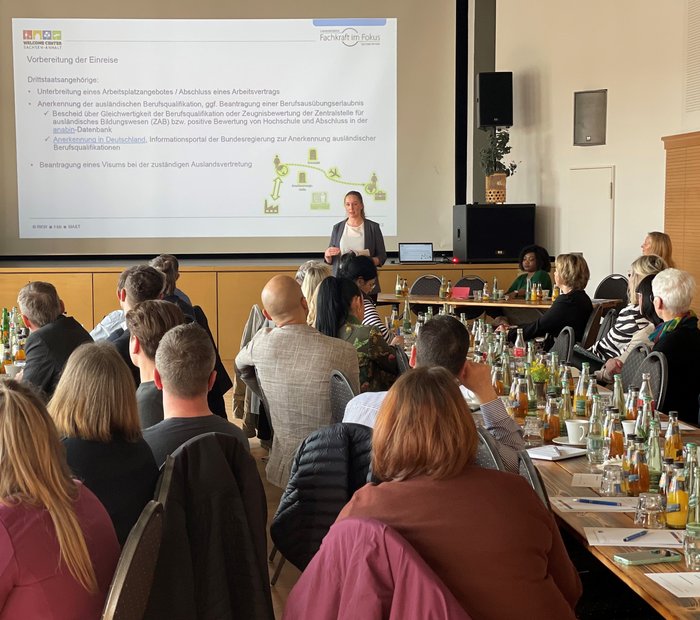 Blick von hinten auf einen Vortrag einer Referentin mit PowerPoint-Präsentation und Publikum an Tischen mit Getränken