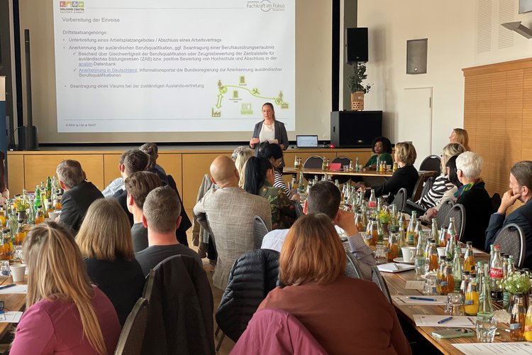 Blick von hinten auf einen Vortrag einer Referentin mit PowerPoint-Präsentation und Publikum an Tischen mit Getränken